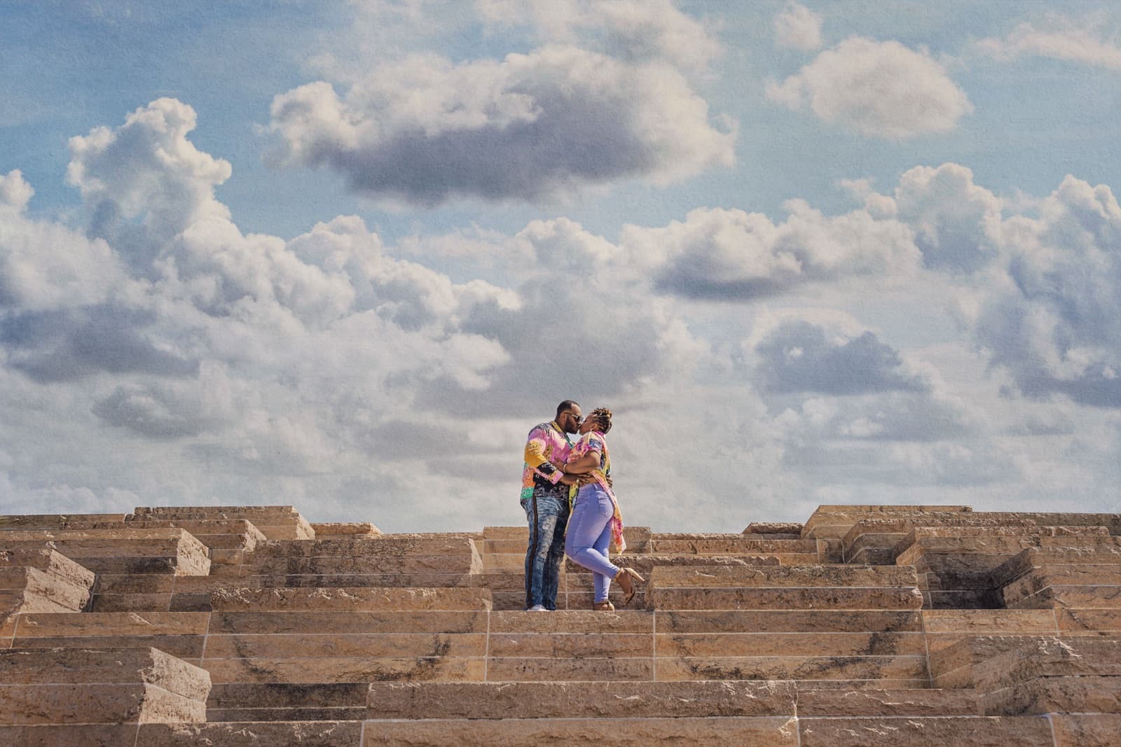 Engaged couple embracing atop sunlit stone steps under a bright blue St. Louis sky