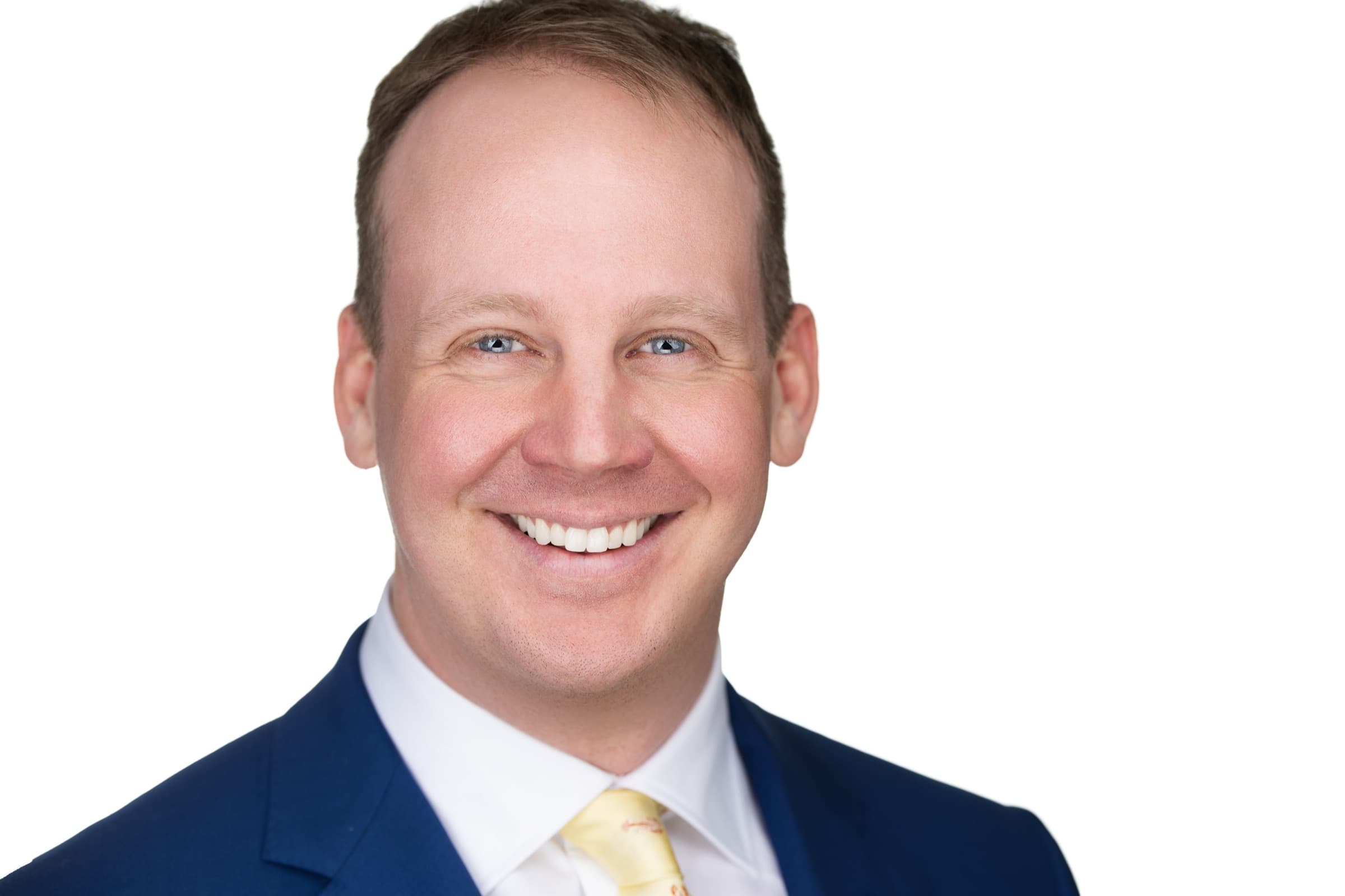 Professional headshot of a smiling man in a navy suit and yellow tie on a white background in St. Louis by Zach Dalin
