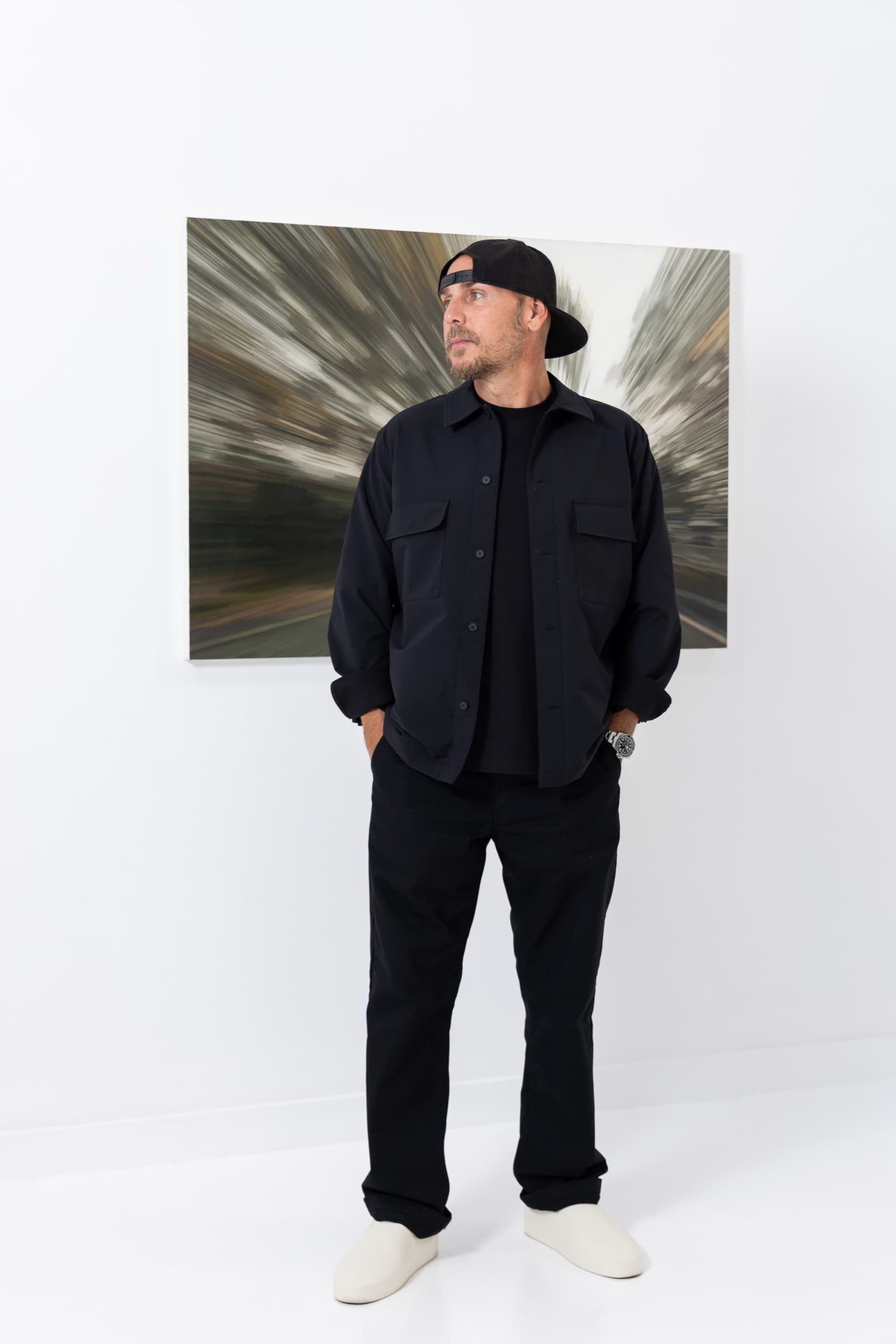 Artist in a backwards cap standing in front of his large abstract painting on a white gallery wall in his St. Louis studio